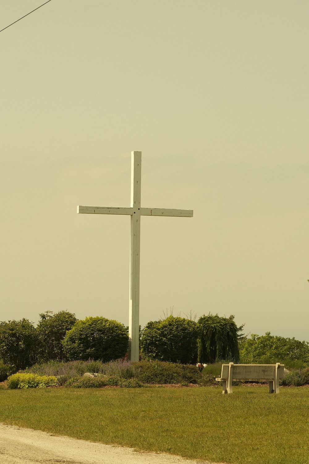 A tall white cross stands on a grassy hill with trees in the background and a wooden bench nearby.