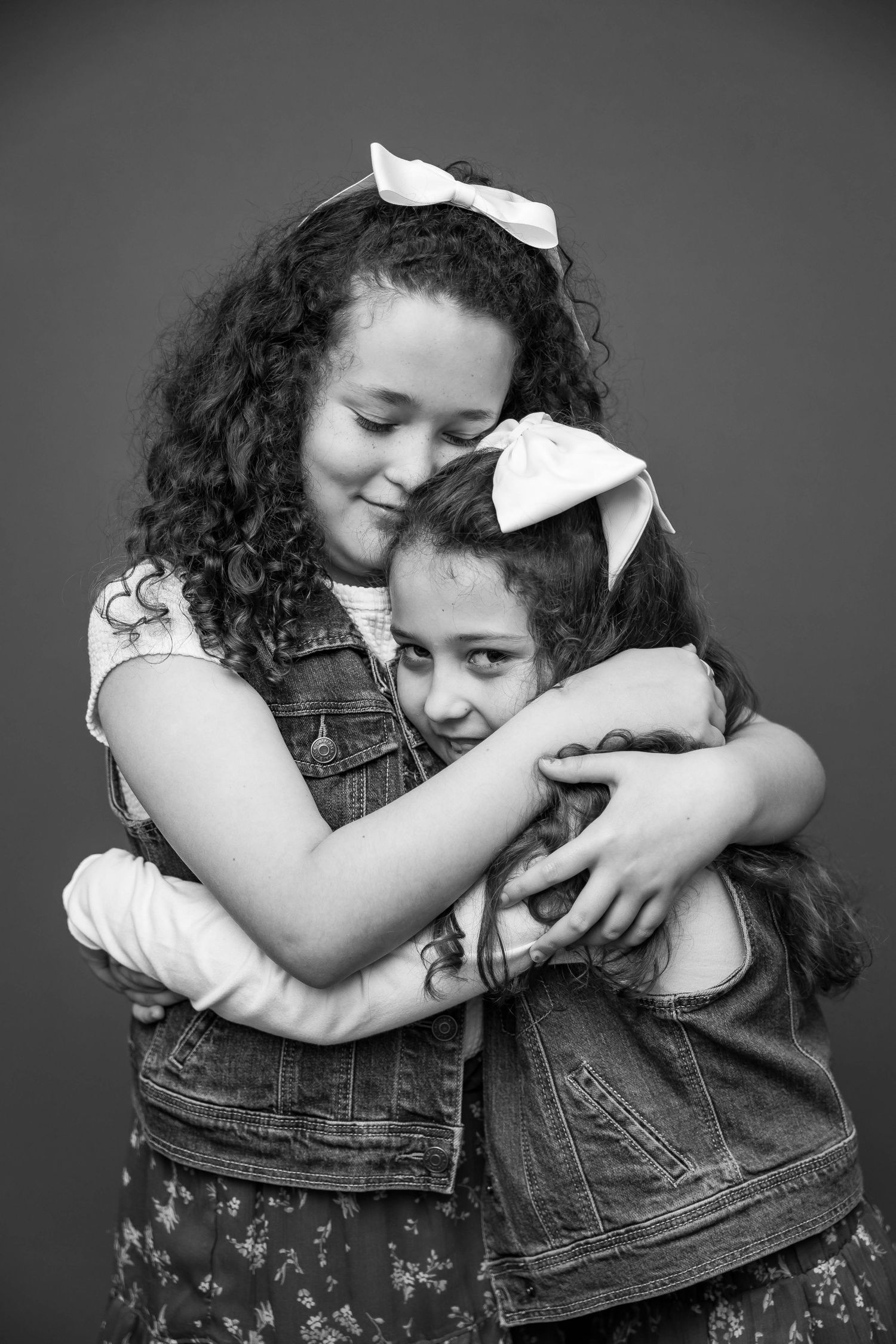 A heartwarming black and white portrait of two young girls wearing matching denim and white bows hugging each other.