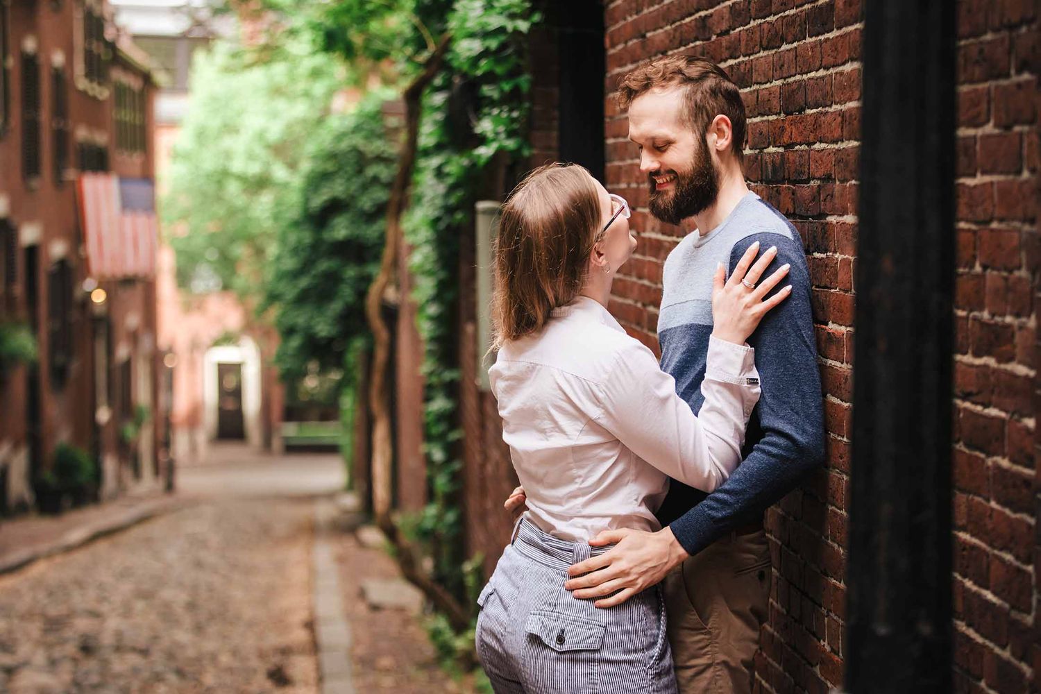 A couple cuddles and smiles affectionately at each other on Acorn Street, surrounded by vibrant green ivy and charming Boston architecture