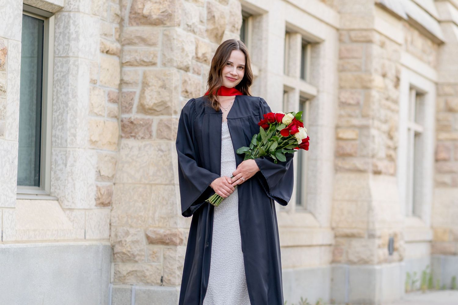 Graduate in black cap and gown holds red roses while standing outside stone building.