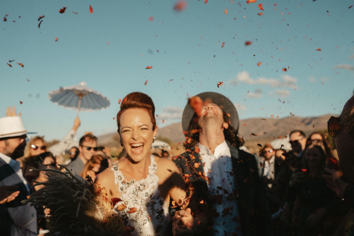A group of wedding attendees celebrate joyfully outdoors with raised bouquets against a backdrop of mountains and trees.