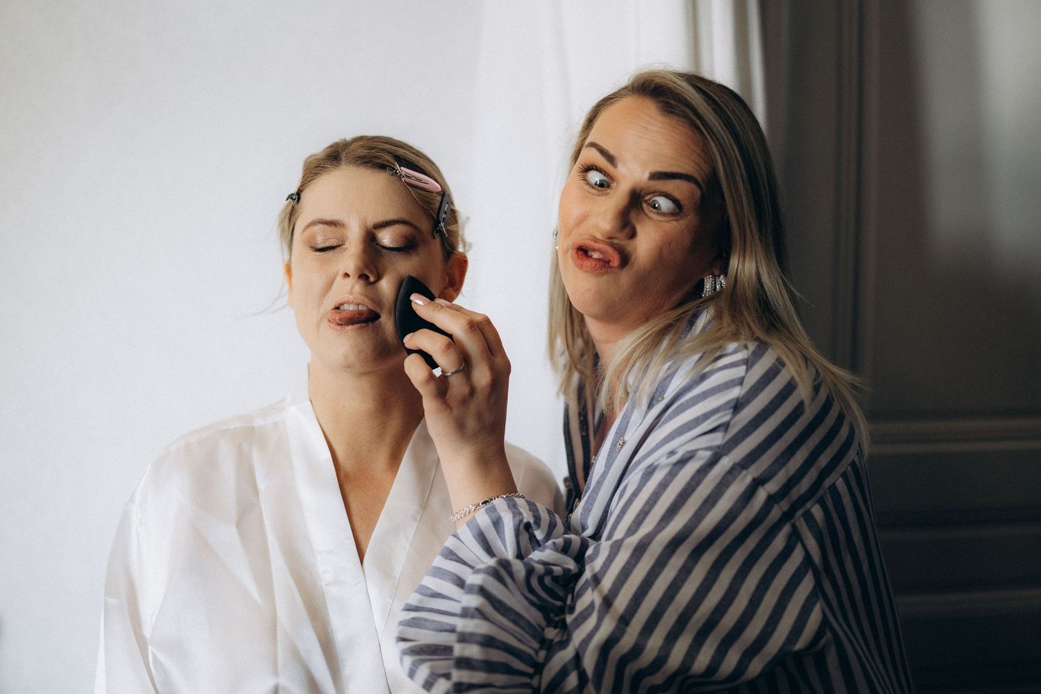 Two people in bathrobes getting ready in morning light by window, one applying lipstick while other watches.