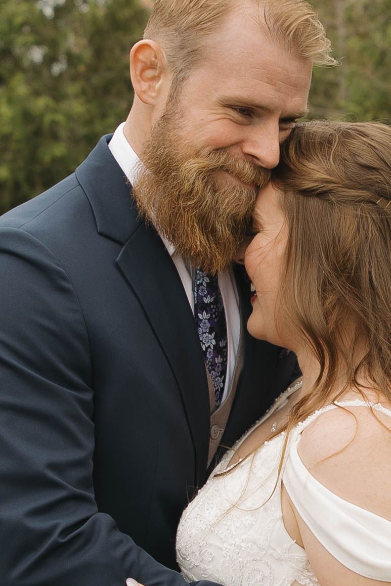 Intimate wedding portrait of a suited groom with ginger beard tenderly embracing his bride in an outdoor setting.
