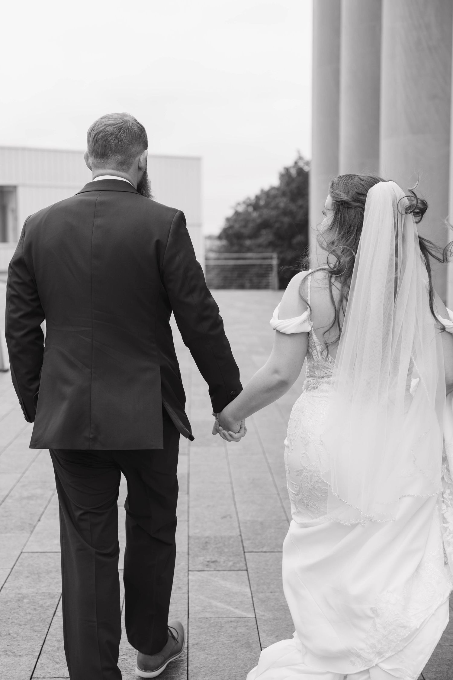 A bride and groom hold hands while walking together on their wedding day, captured in black and white in downtown Kansas City.