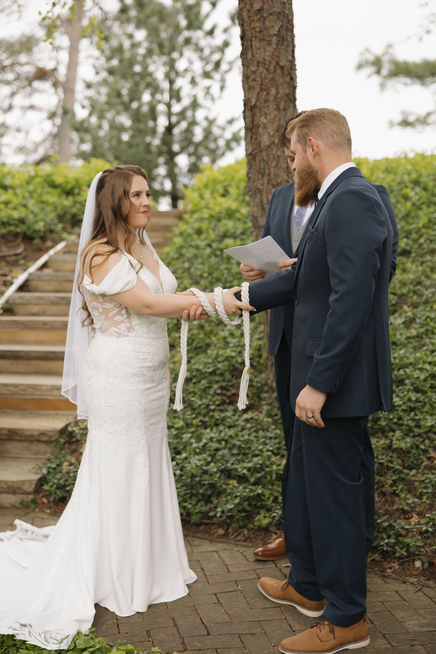Couple exchanges vows during outdoor wedding ceremony on brick path surrounded by greenery and steps.
