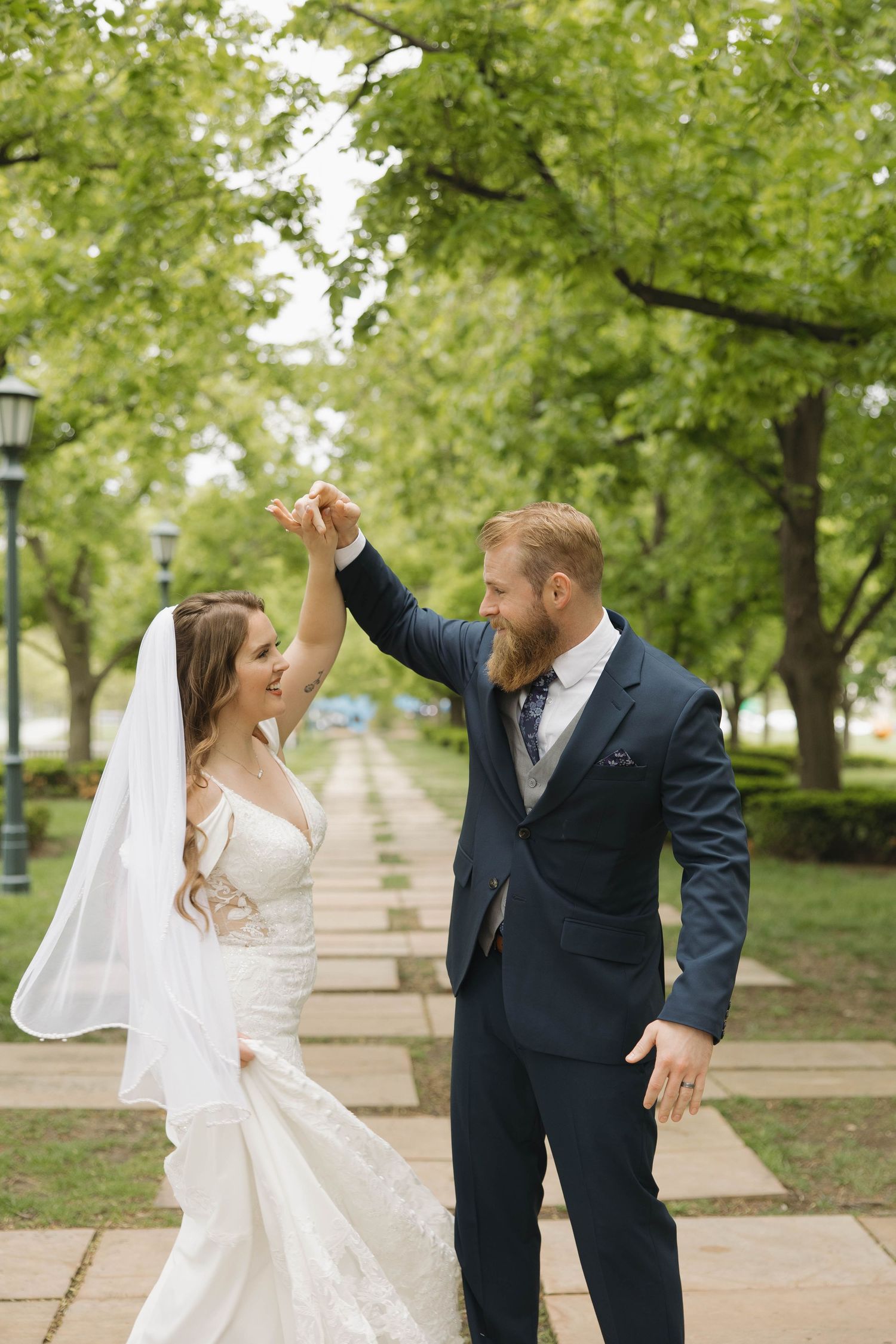 Newlyweds dance and celebrate on a tree-lined path with classic lamp posts and greenery.