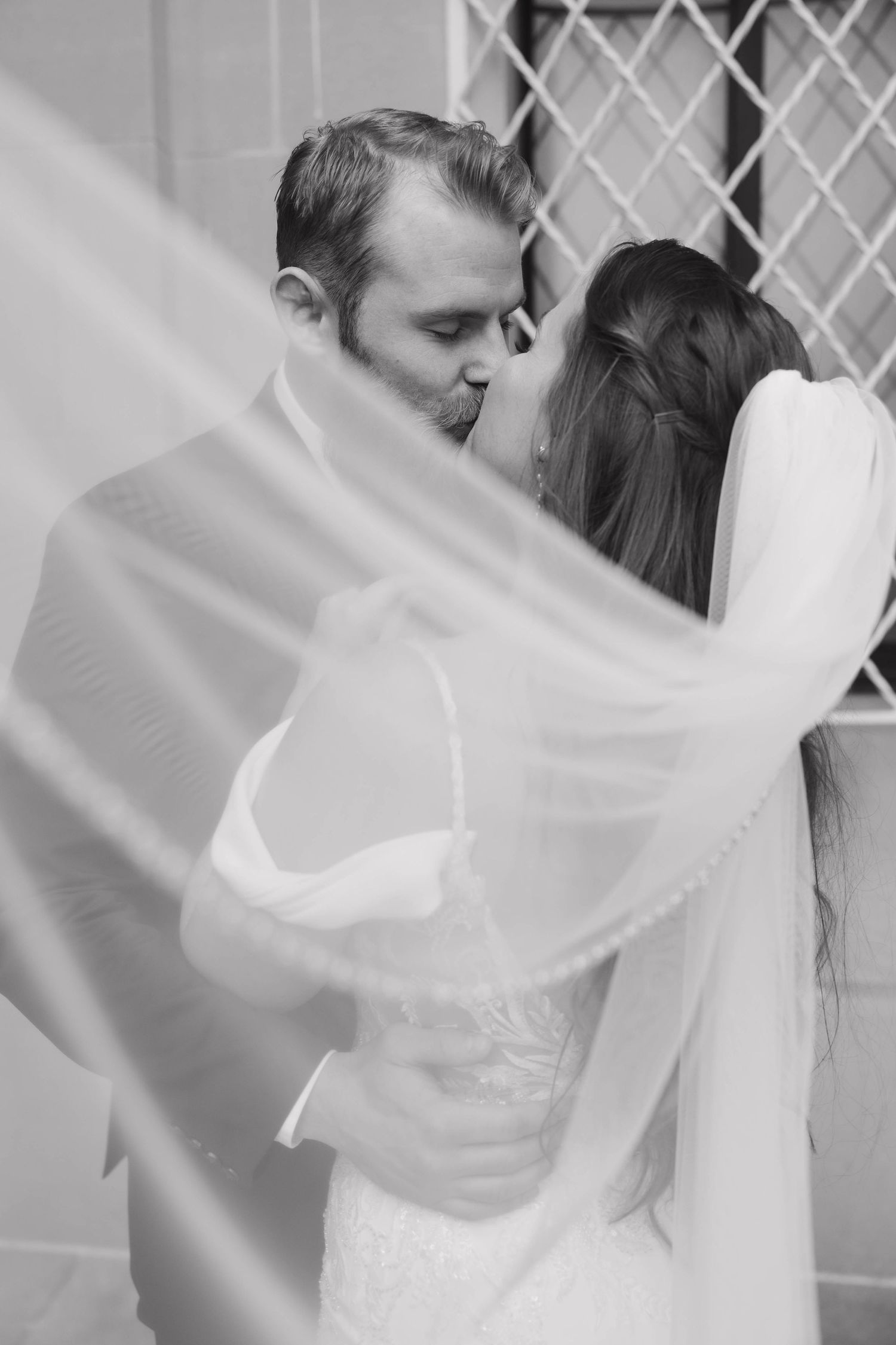 Black and white wedding photo capturing romantic kiss through flowing white veil.