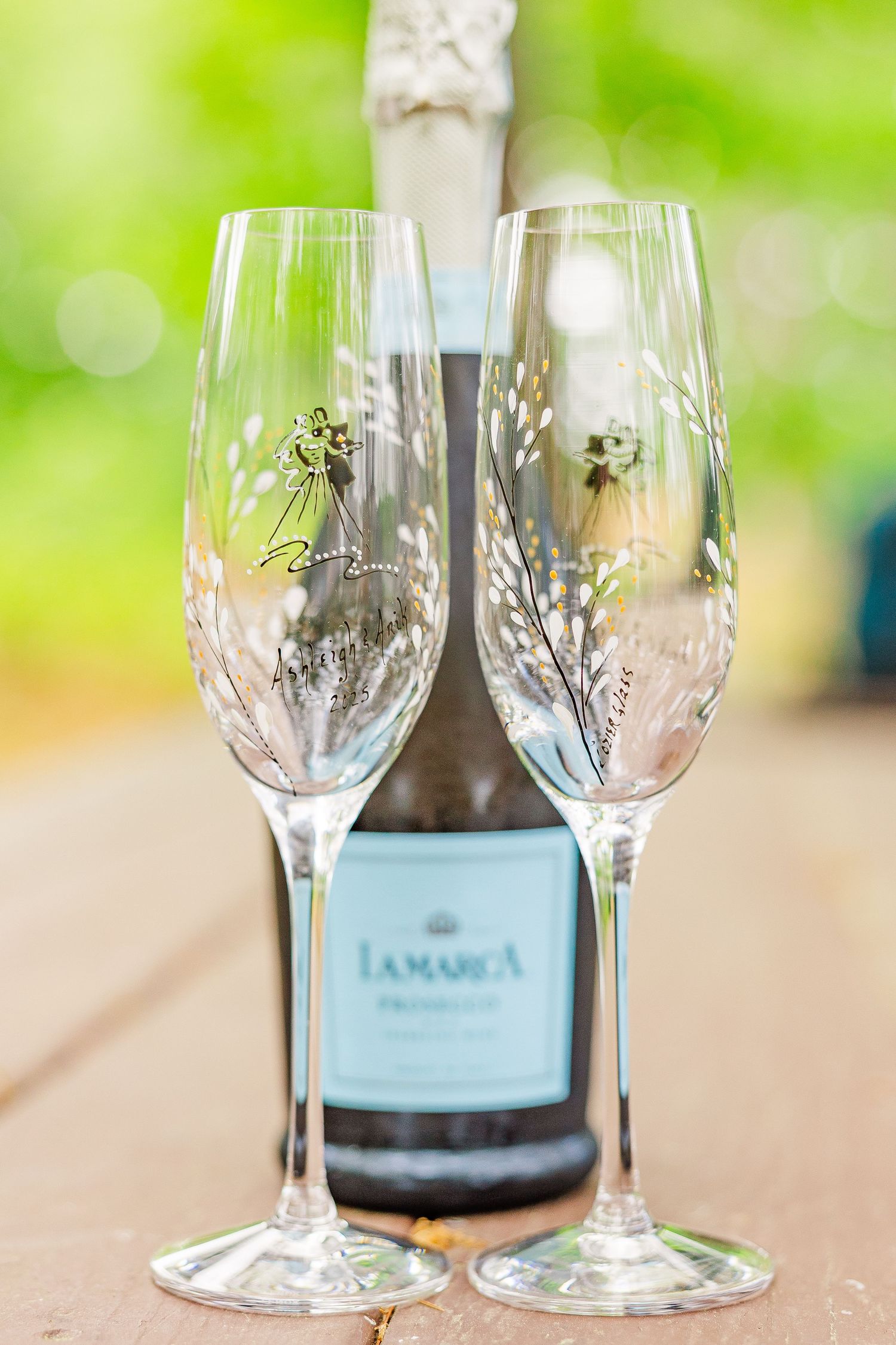 Two elegant champagne flutes with delicate floral designs stand in front of a La Marca prosecco bottle against a blurred green background.