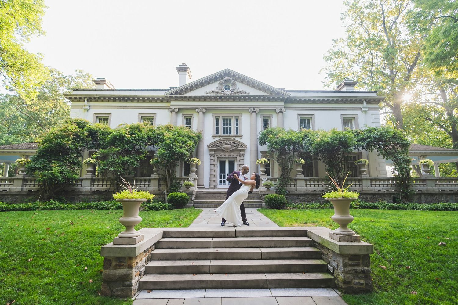 Elegant white mansion with neoclassical columns and landscaped garden features stone steps leading to main entrance.