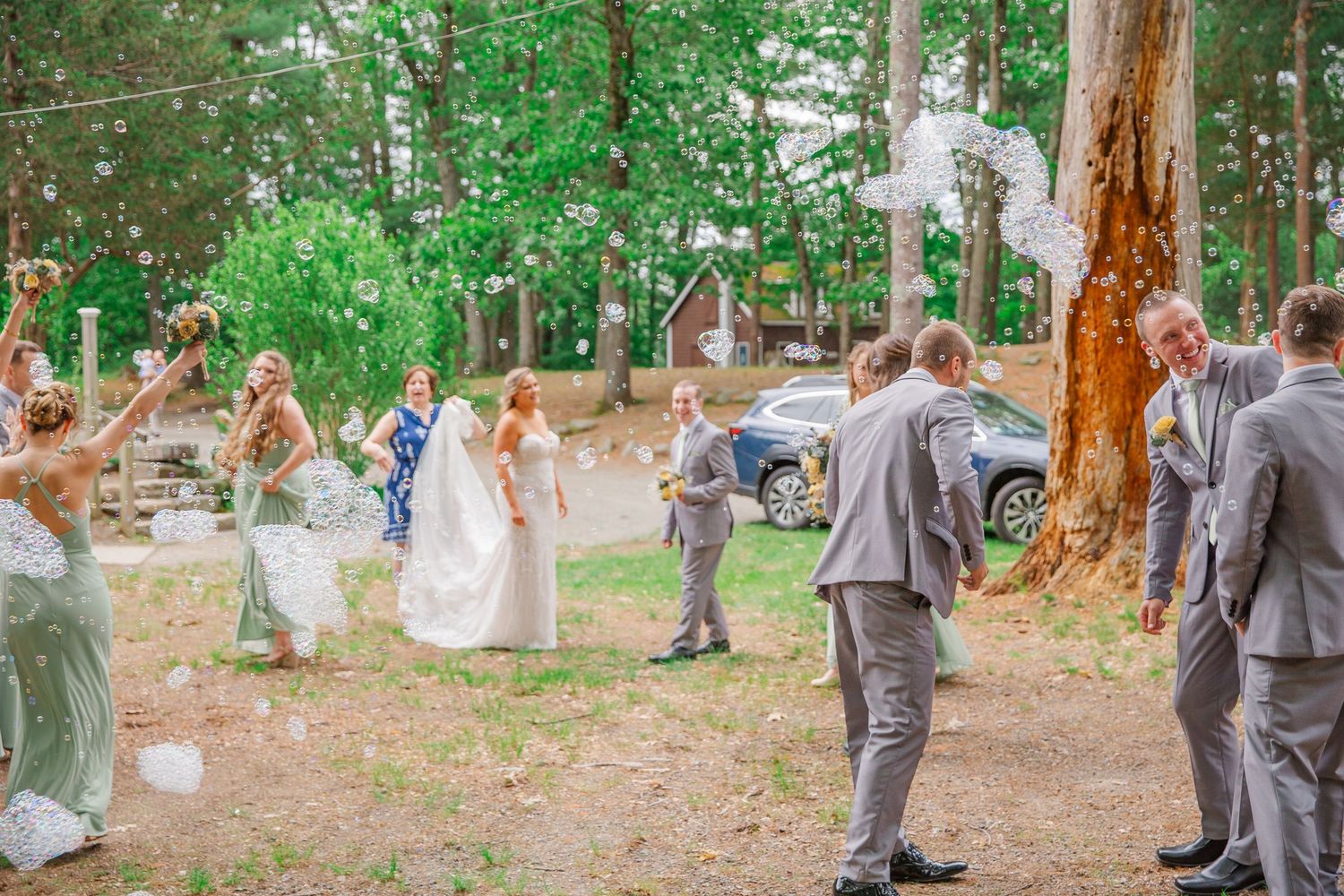 Wedding party celebrates outdoors in a wooded setting with guests dancing and having fun on a sunny day.