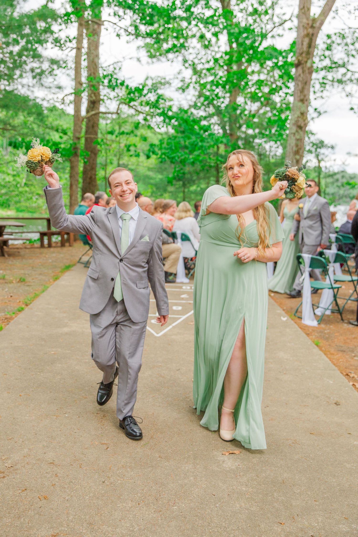 Wedding party members in sage green dresses and gray suits striking fun dance poses during an outdoor celebration.