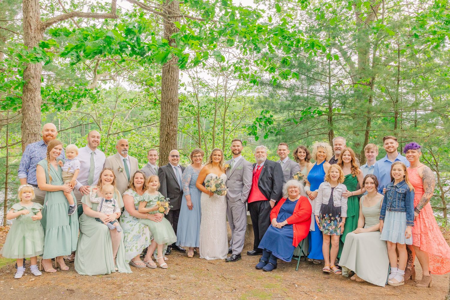A large group in formal attire poses together outdoors in a wooded setting during a special celebration.