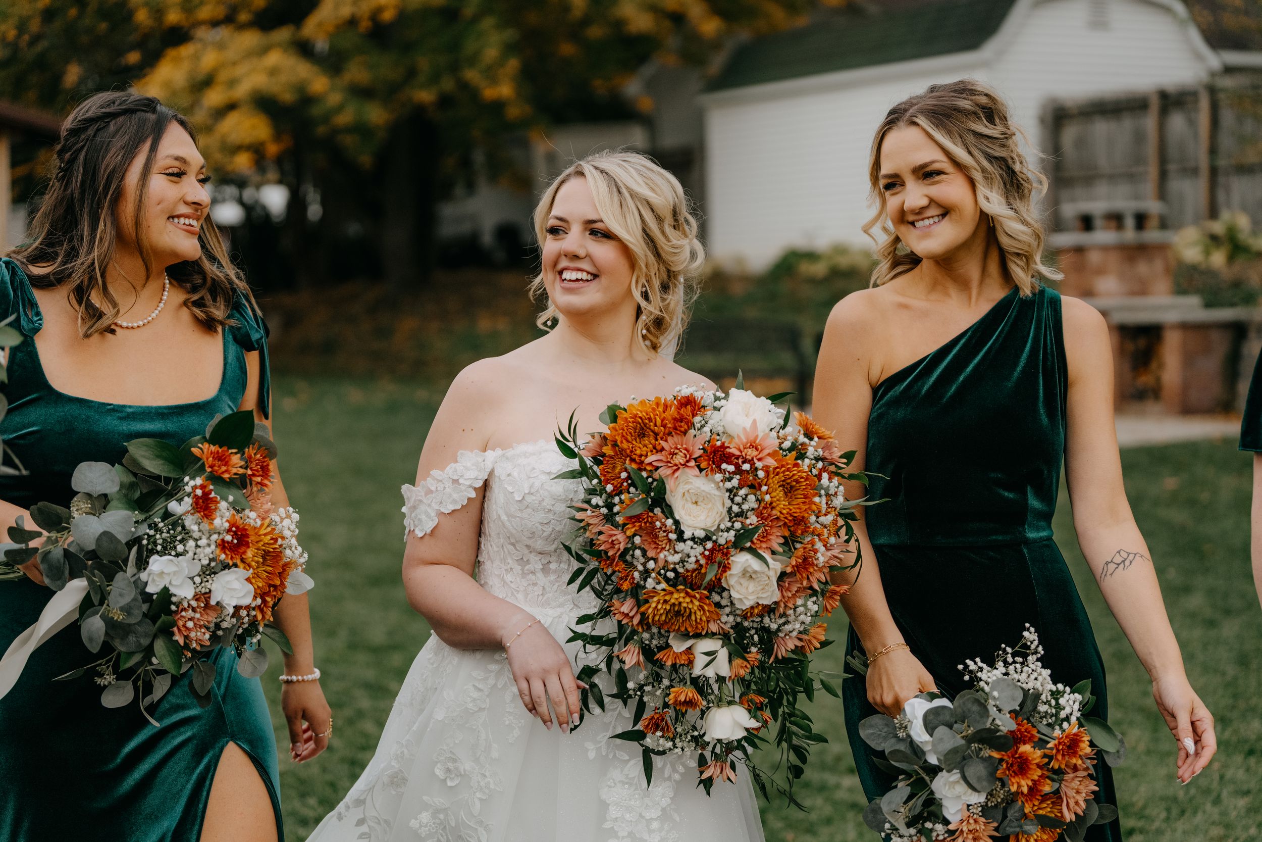 Bridesmaids in coordinating emerald green and blush dresses hold orange and rust-colored fall bouquets at outdoor wedding.