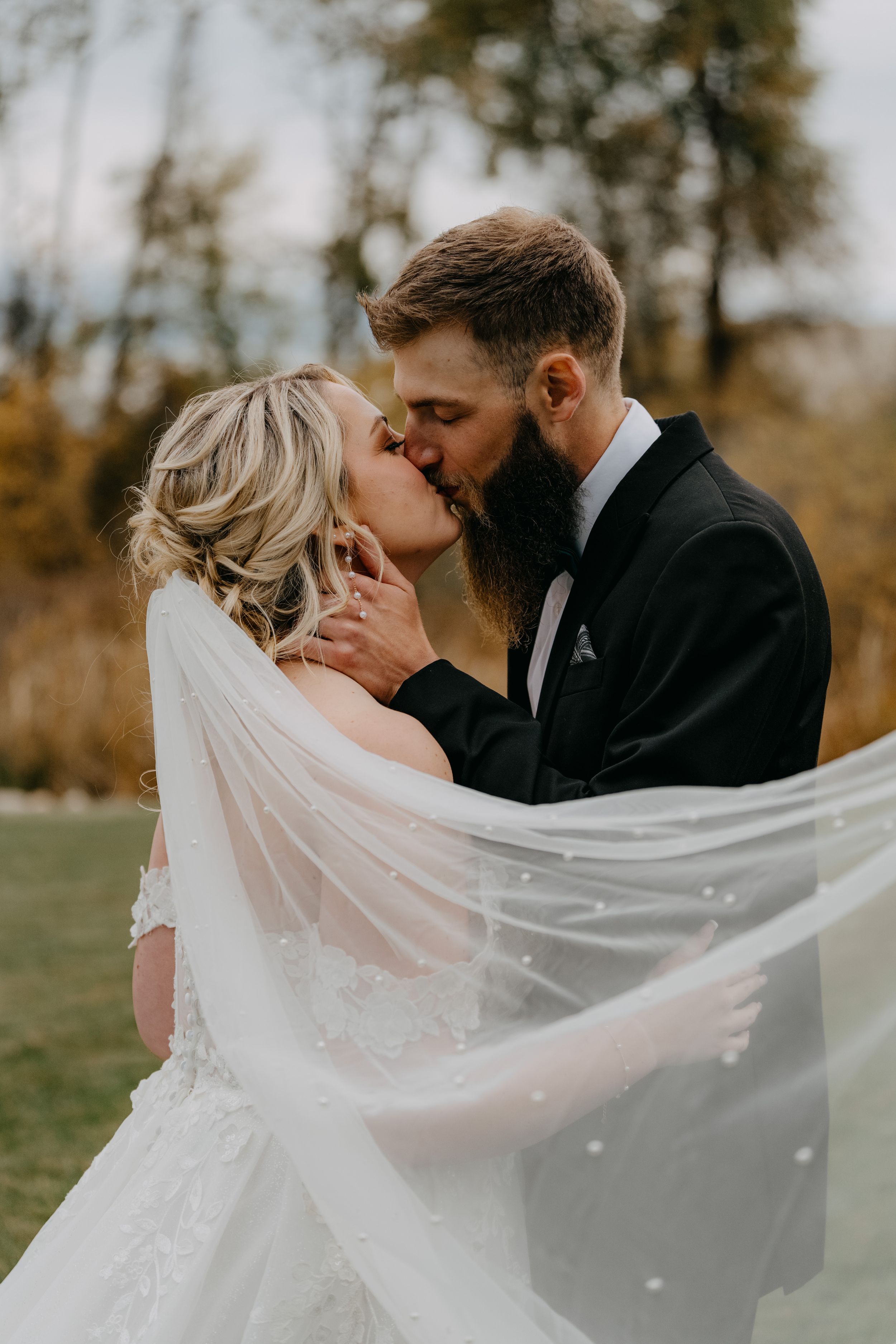 Romantic wedding couple sharing intimate kiss outdoors while bride's lace veil flows in autumn breeze.
