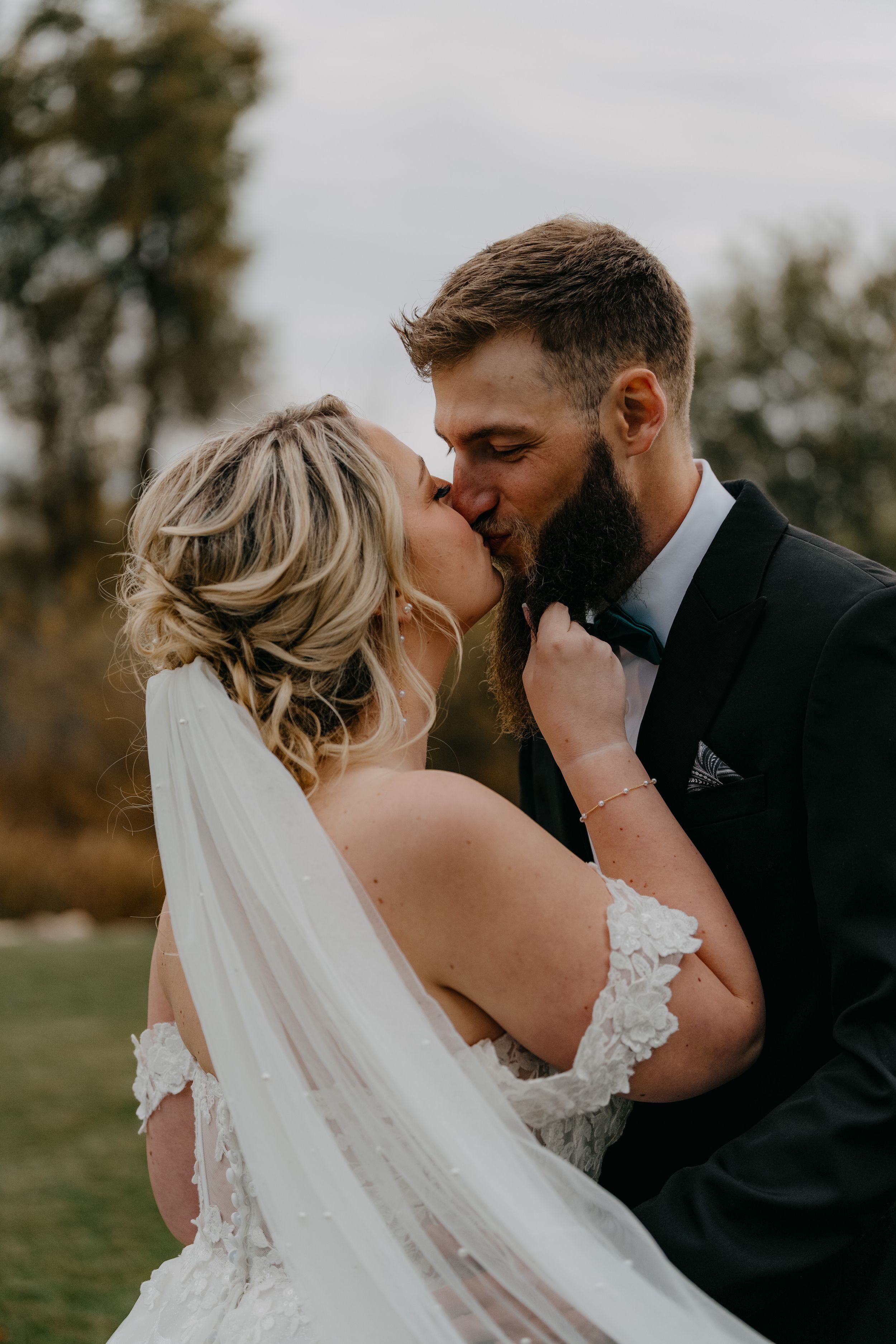 Wedding couple share romantic kiss outdoors with bride in lace gown and long veil and groom in black suit.