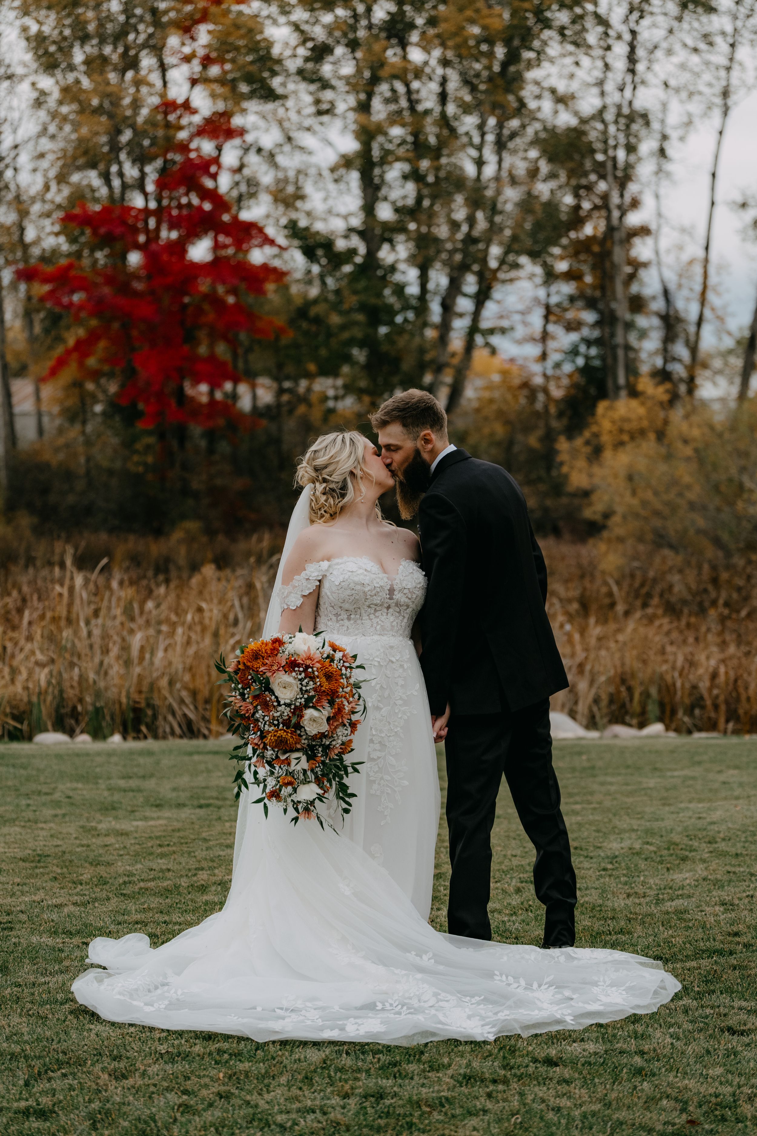 Newlywed couple embracing in an autumn field with red maple trees and tall grasses in background at sunset.