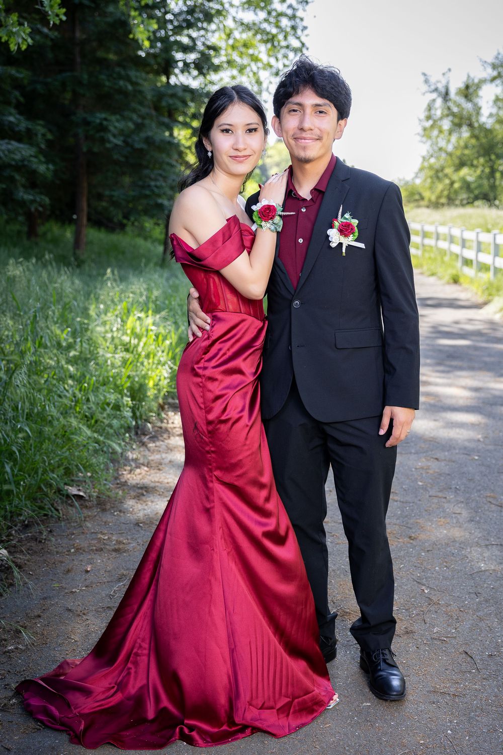 A couple in formal attire poses together outdoors, with a red dress and black suit against a natural background.