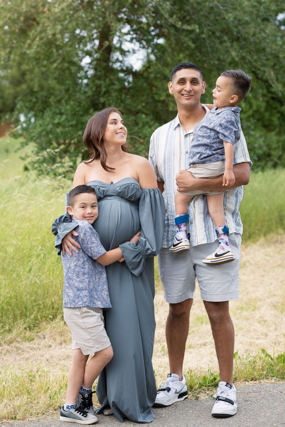 A family stands together outdoors in a grassy area, with a pregnant woman in a long gray dress and children in matching outfits.