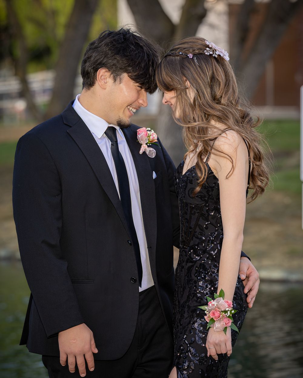 A smiling couple in prom attire, man in black suit with boutonniere, woman in black sequin gown with floral corsage.