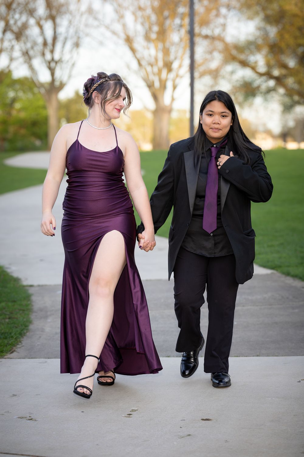 Couple in formal prom attire walking outdoors, woman in purple gown and man in black tuxedo with purple tie.