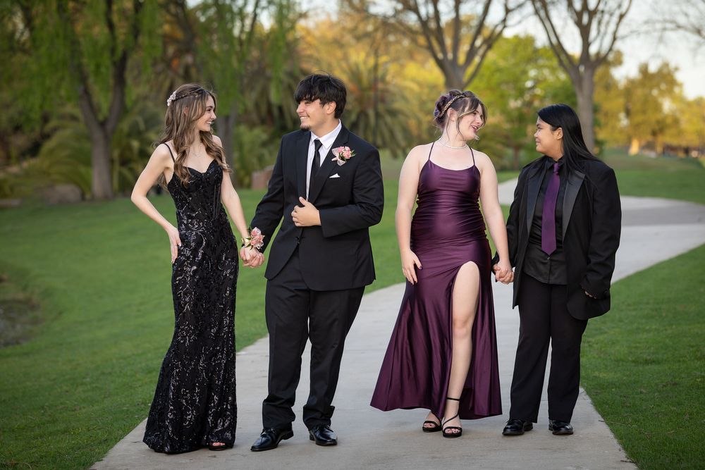 Four teenagers in formal prom attire walk together outdoors on a path surrounded by lush green trees.