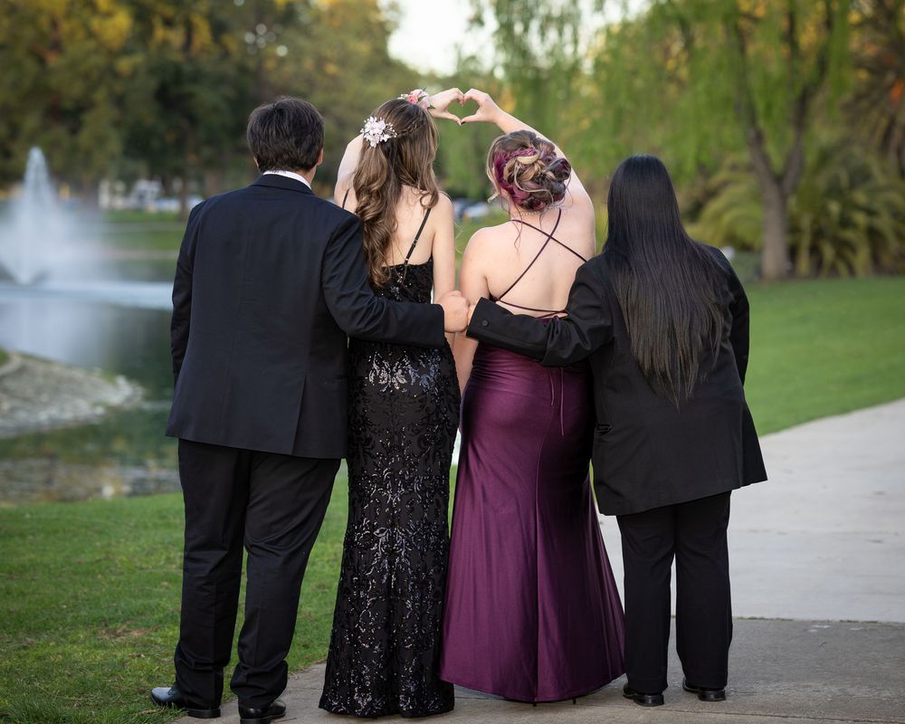 Four friends in formal attire pose together from behind at an outdoor park near a fountain.
