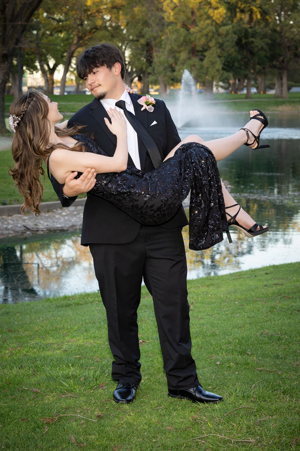 Young couple in formal wear poses by a fountain, man in tuxedo carrying woman in black sequin dress at prom.