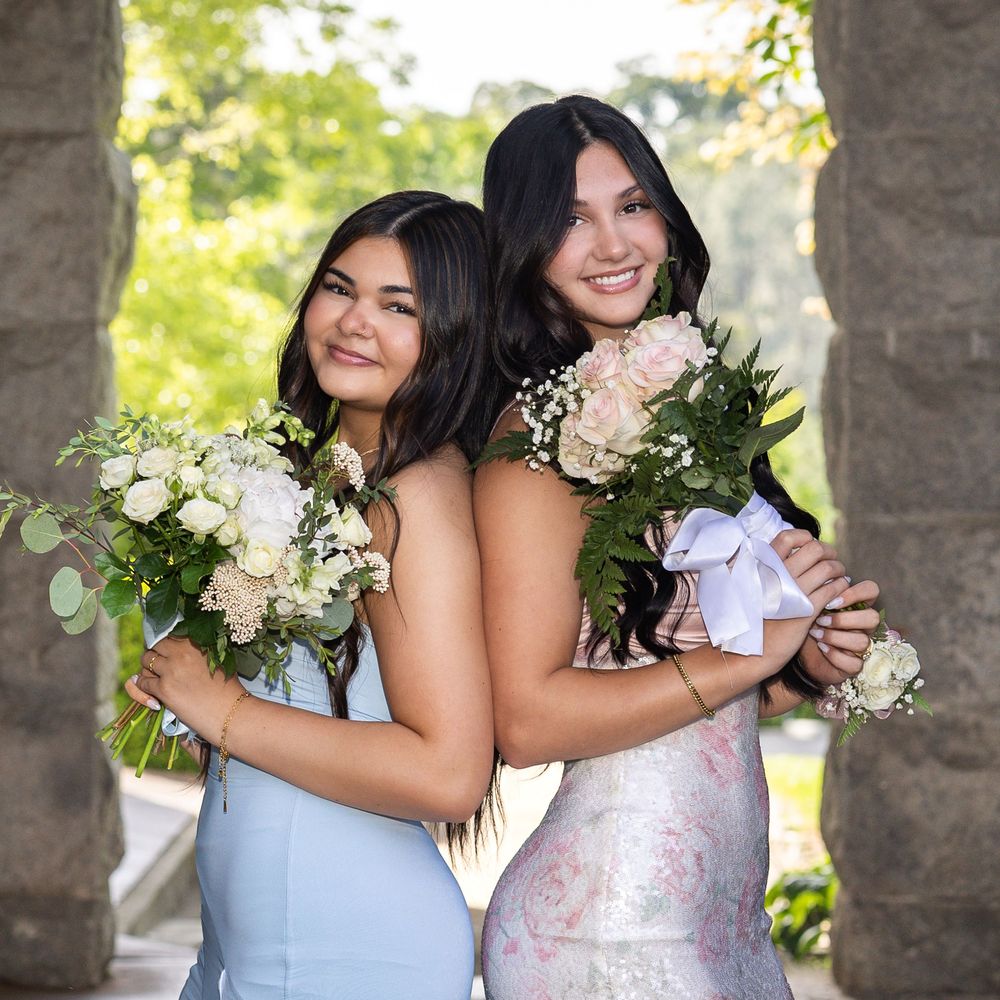 Two young women in formal dresses holding white floral bouquets pose back-to-back under a stone arch outdoors.