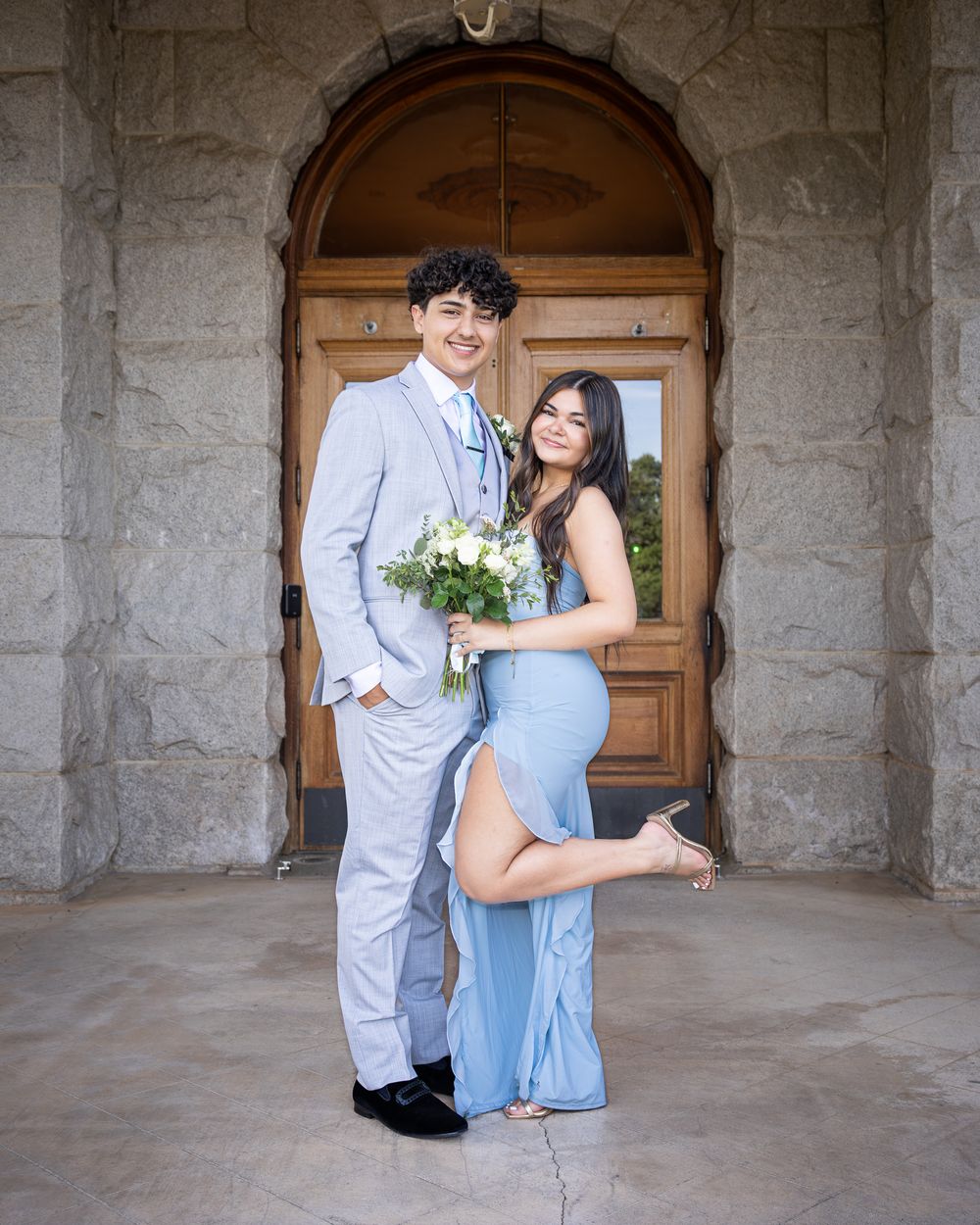A couple in matching light blue prom attire poses in front of elegant wooden doors, holding a white floral bouquet.