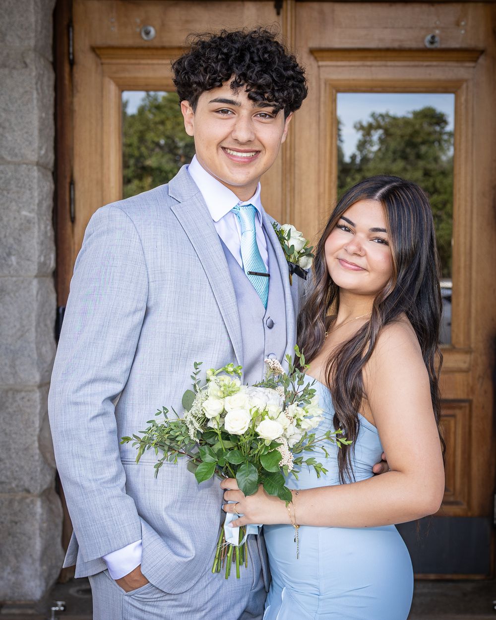 Young couple in coordinating light blue prom attire, holding white floral bouquet, posing in front of wooden doors.