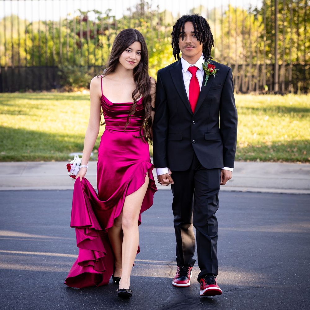 A couple dressed in prom attire, girl in red gown and boy in black suit with red tie, walking hand in hand.