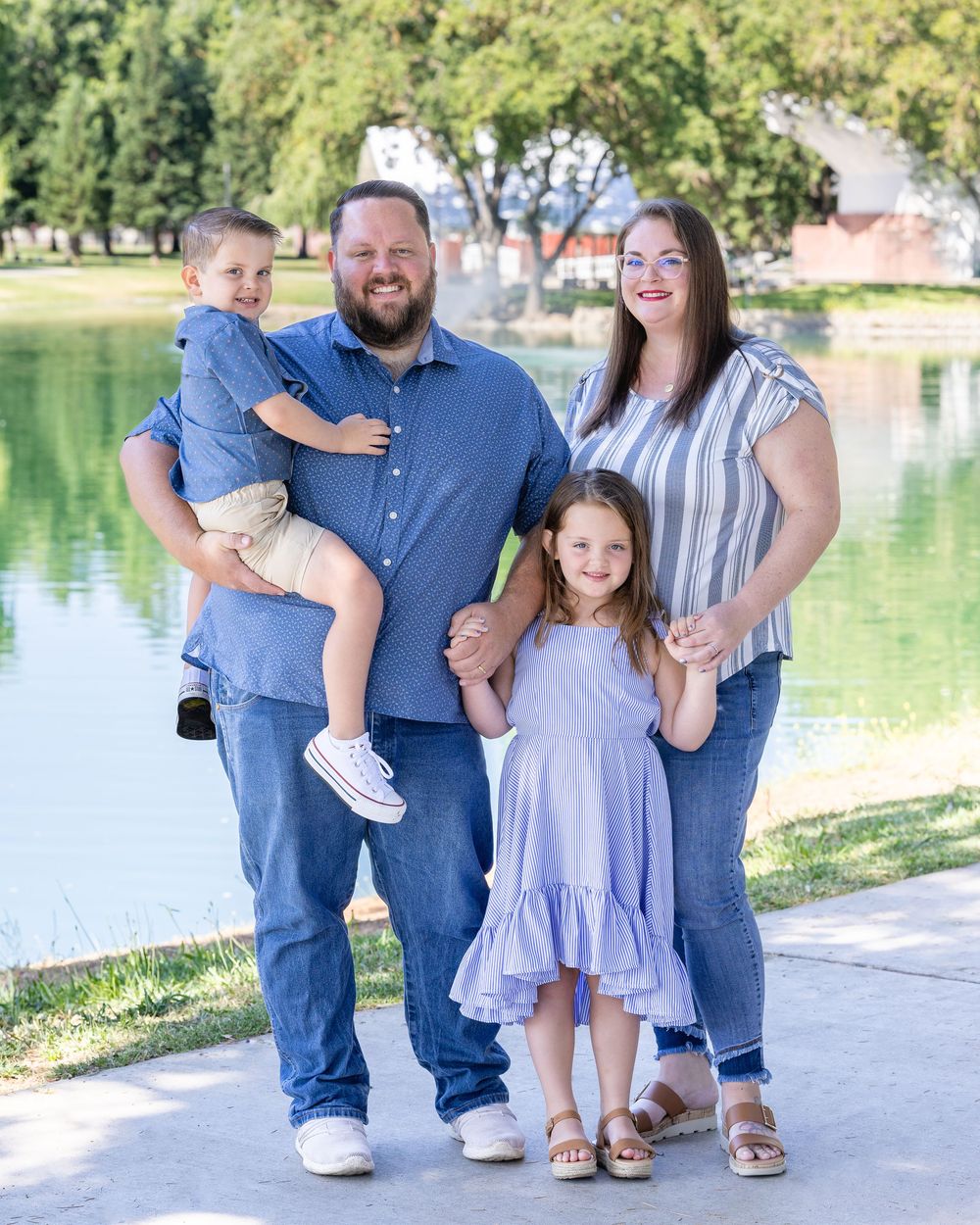 A family stands together by a tranquil pond on a sunny day, wearing casual blue and purple outfits.