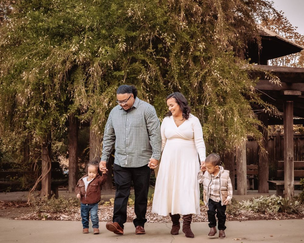 A family walks together holding hands along a pathway under autumn trees near a rustic wooden structure.