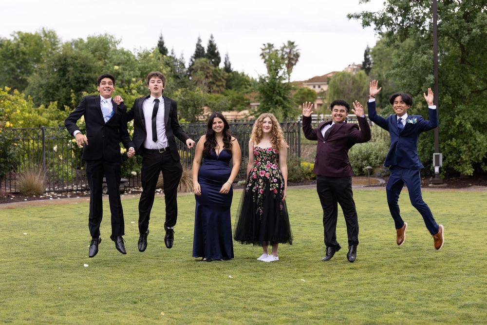 Six teens in formal attire jump joyfully on grass during a prom or formal event outdoors.