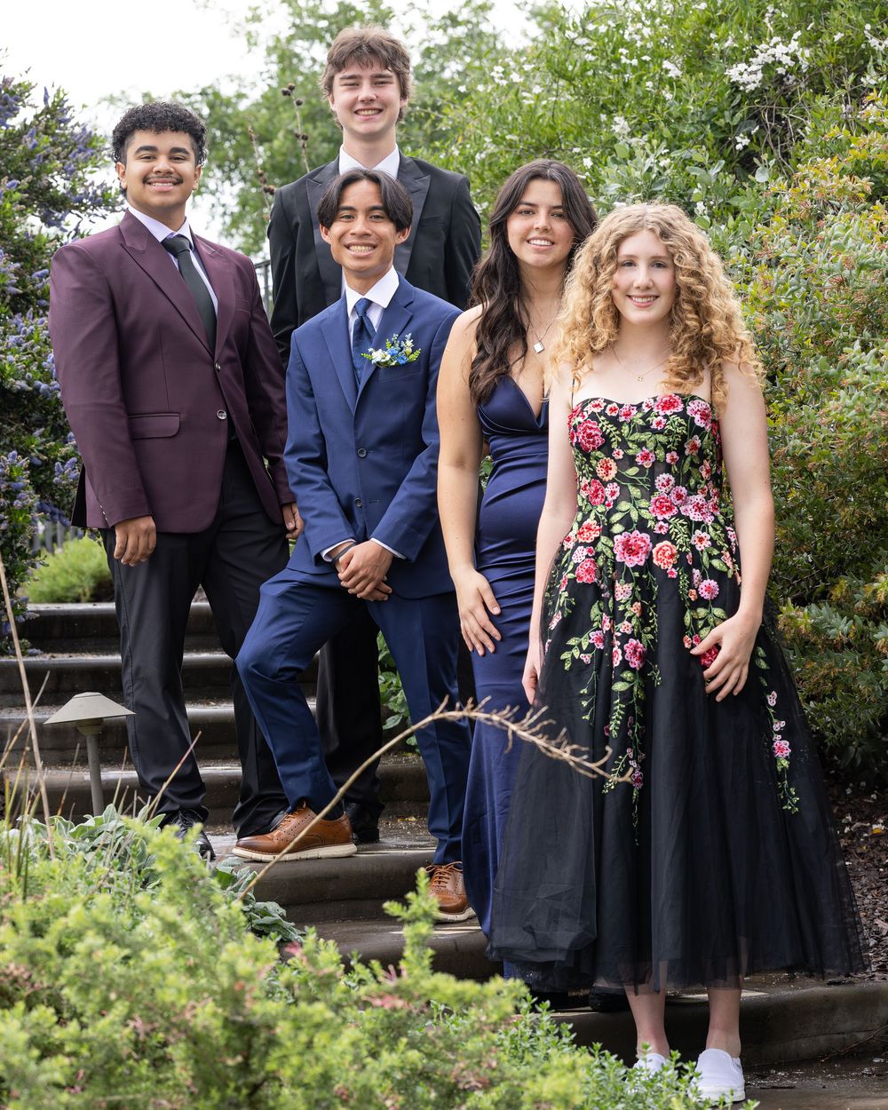 Five teenagers dressed in formal prom attire pose on garden steps surrounded by lush greenery.
