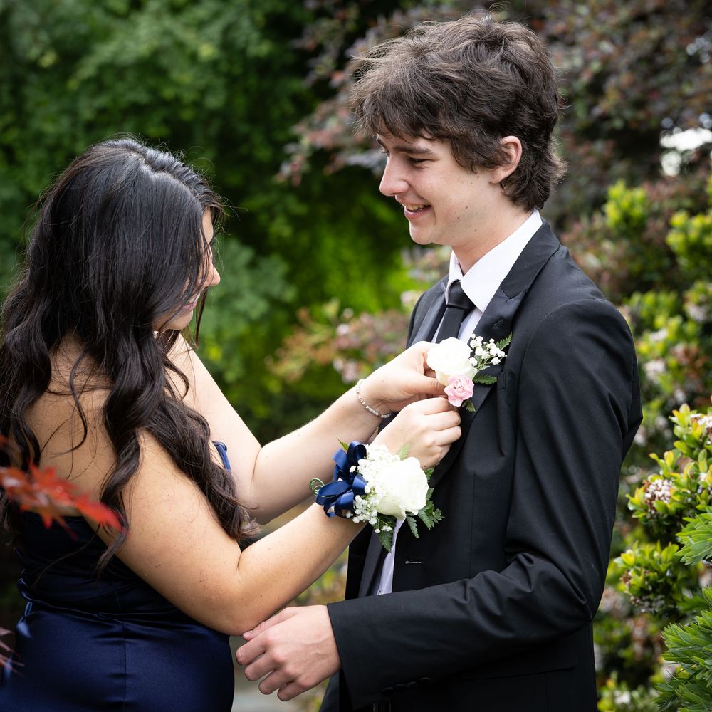 A young couple in formal attire pins a boutonniere before prom, surrounded by lush green garden foliage.
