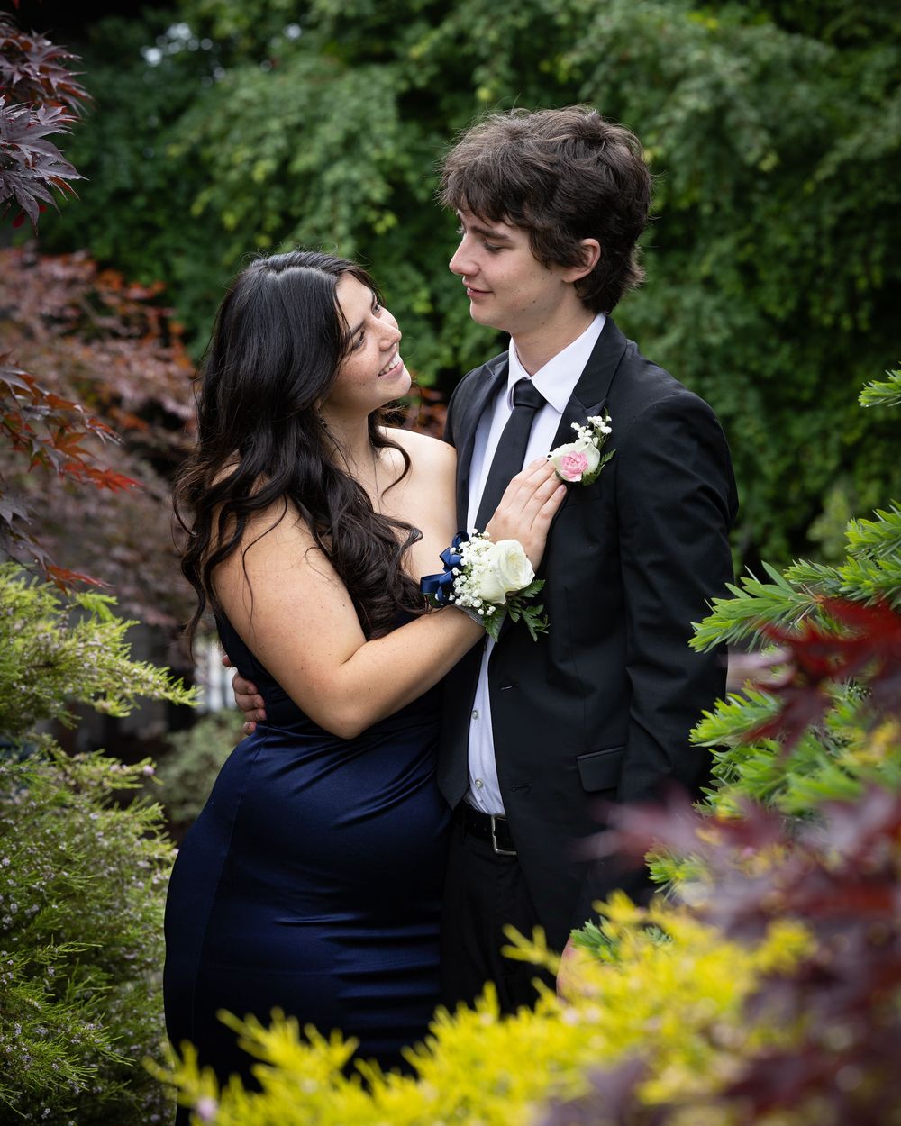 A couple in formal prom attire poses in a garden, the young woman in navy blue touching her date's boutonniere.
