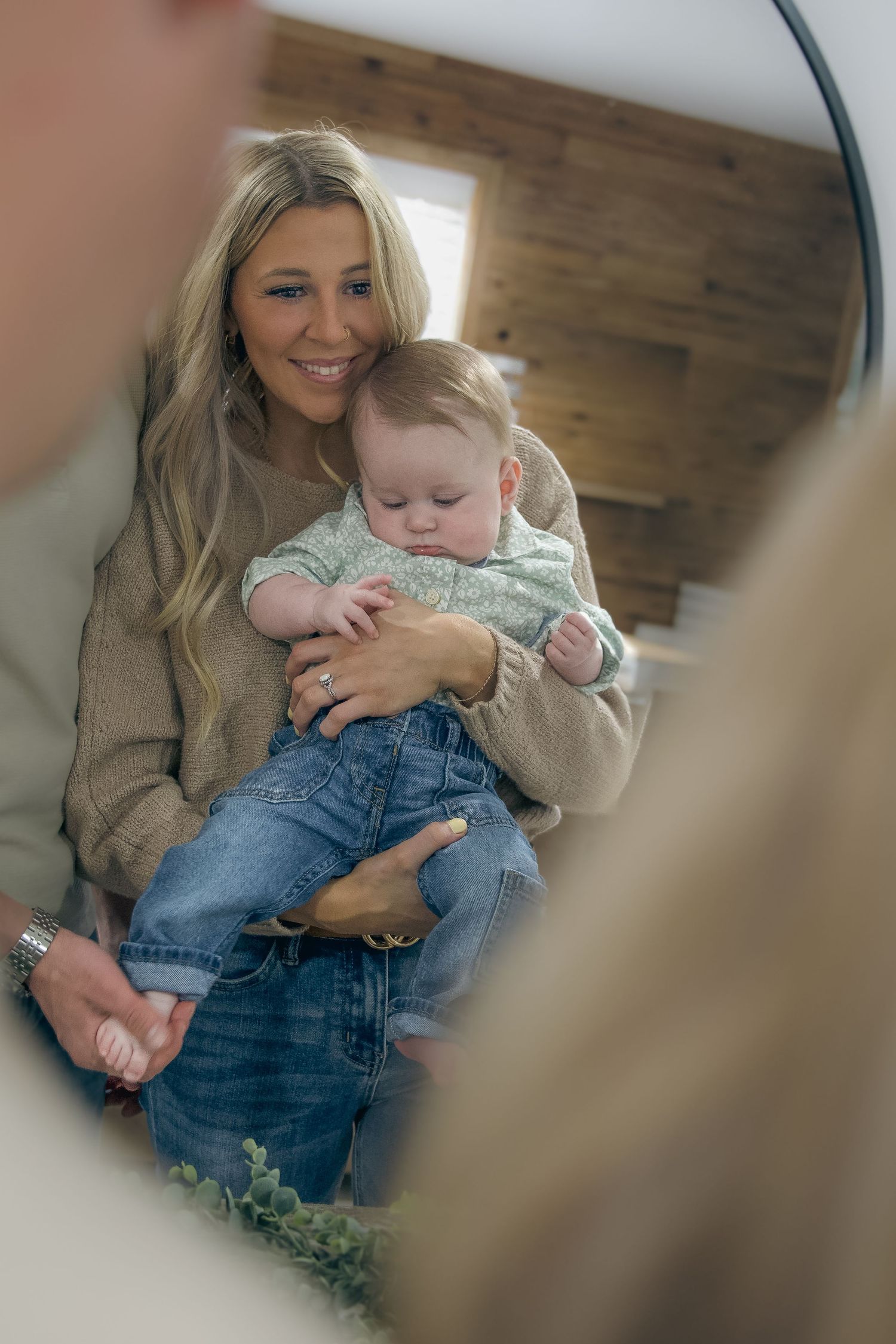 A parent wearing a beige sweater holds a sleepy baby in denim overalls while sitting together, reflected in a mirror.