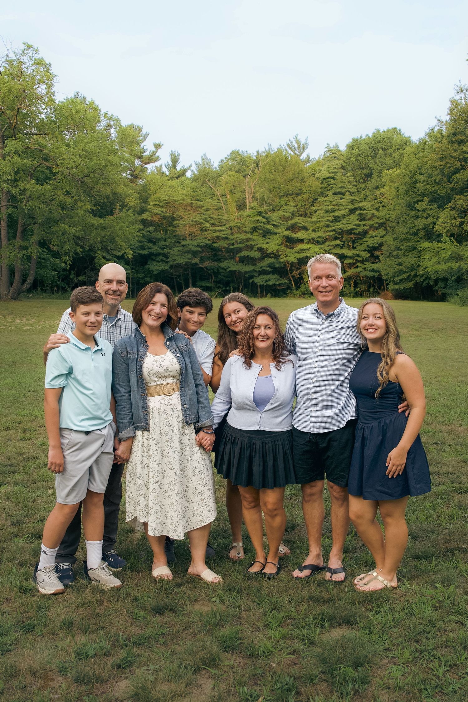 A group of people wearing casual summer attire pose together on grass with trees in the background.