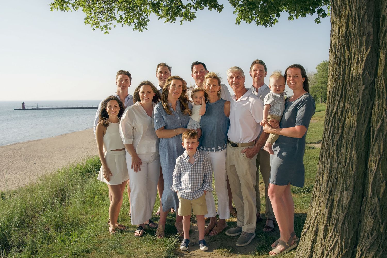 People gathered outdoors near a tree with a waterfront view on a sunny day.