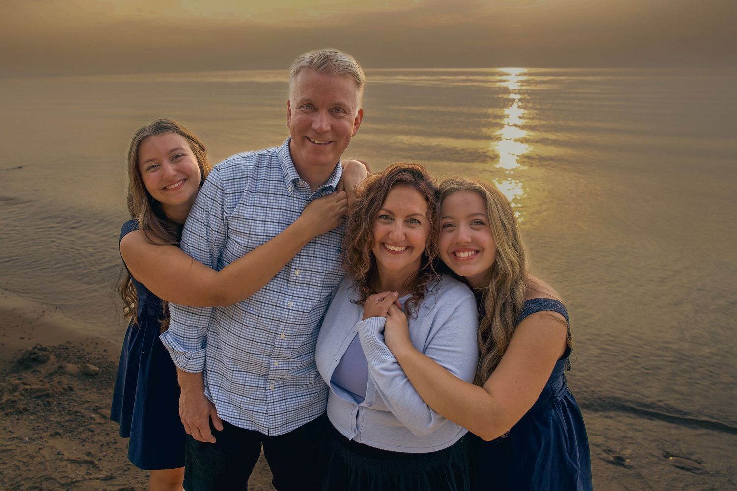 A family poses together on the beach at sunset with golden light reflecting on the ocean in the background.
