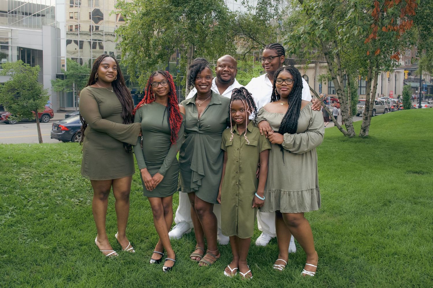 A group stands together smiling on a green lawn in front of buildings, wearing coordinated olive green and neutral outfits.