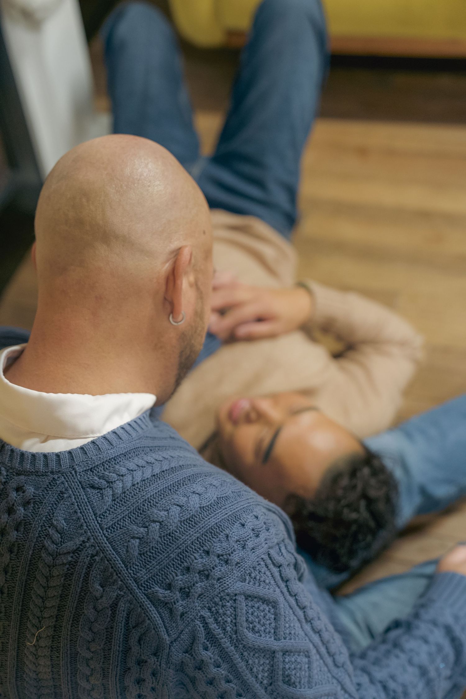 Person wearing a blue cable knit sweater sits near someone reclining on a wooden floor.