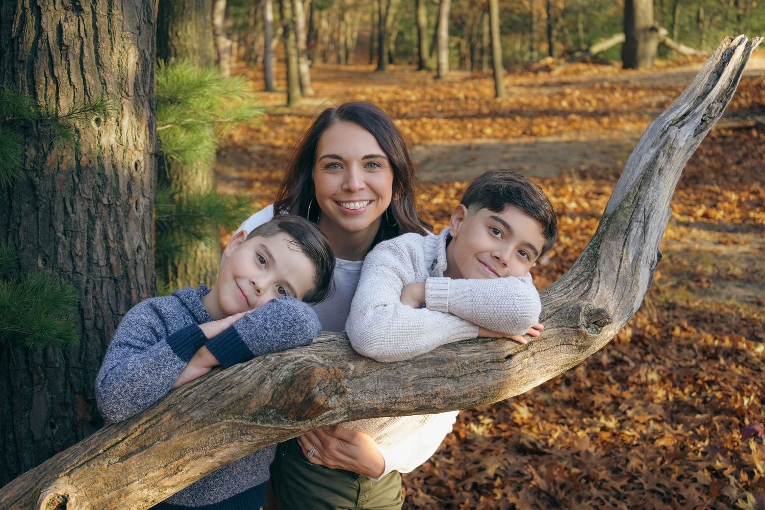 Family members relax and pose together on fallen tree branches in autumn forest setting with orange leaves on ground.