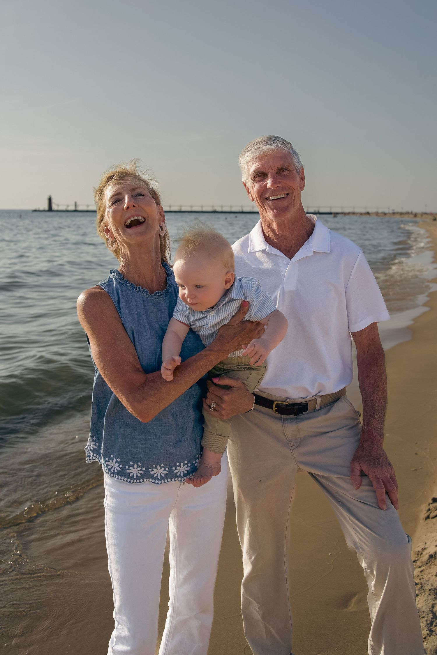 A happy family enjoys a sunset moment together on the beach with waves and a lighthouse in the background.