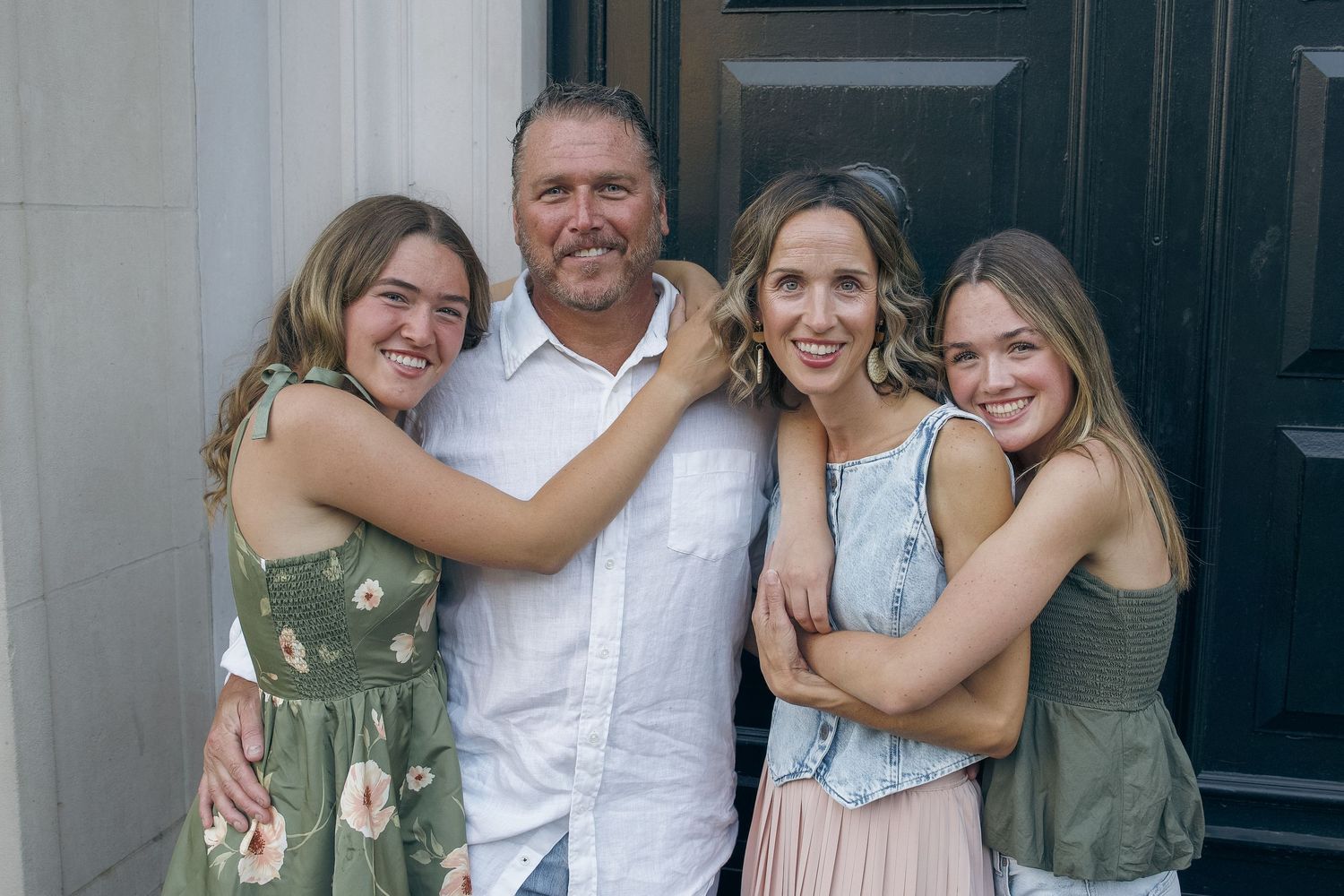 A family of four smiling together outdoors, wearing casual summer attire in front of a dark door.