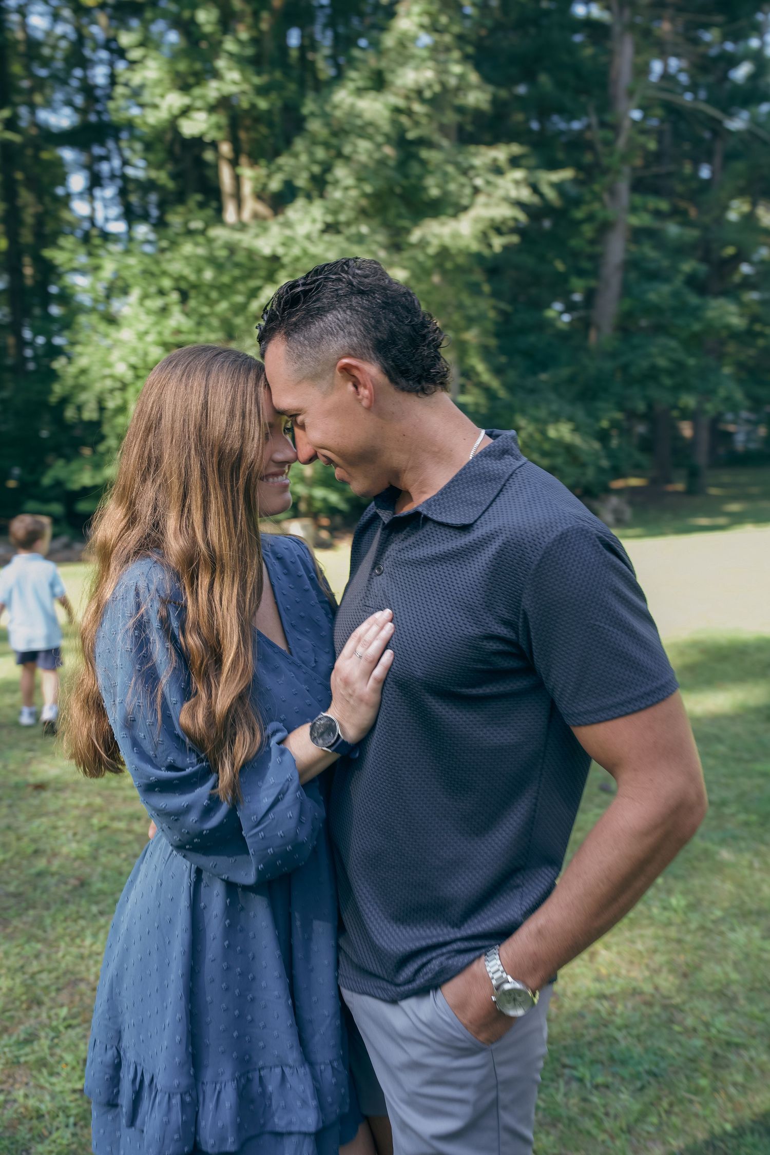 A couple shares an intimate moment together in a park setting with green trees in the background.