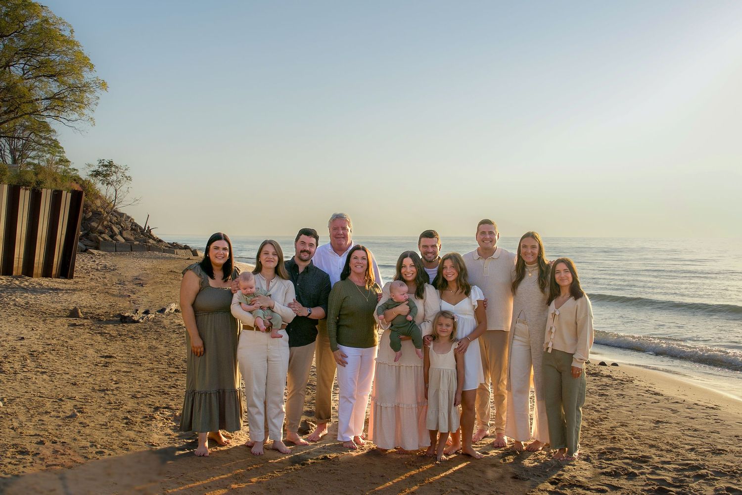 Group of people wearing light colored clothing gathered on a beach at sunset for family photos.