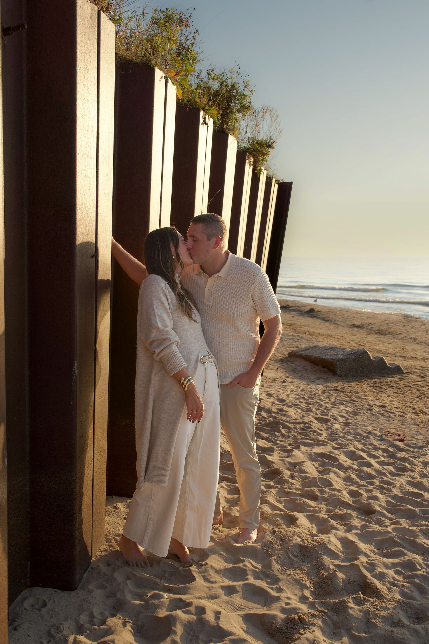 Person in white dress walks along sandy beach with wooden pillars casting shadows at sunset.