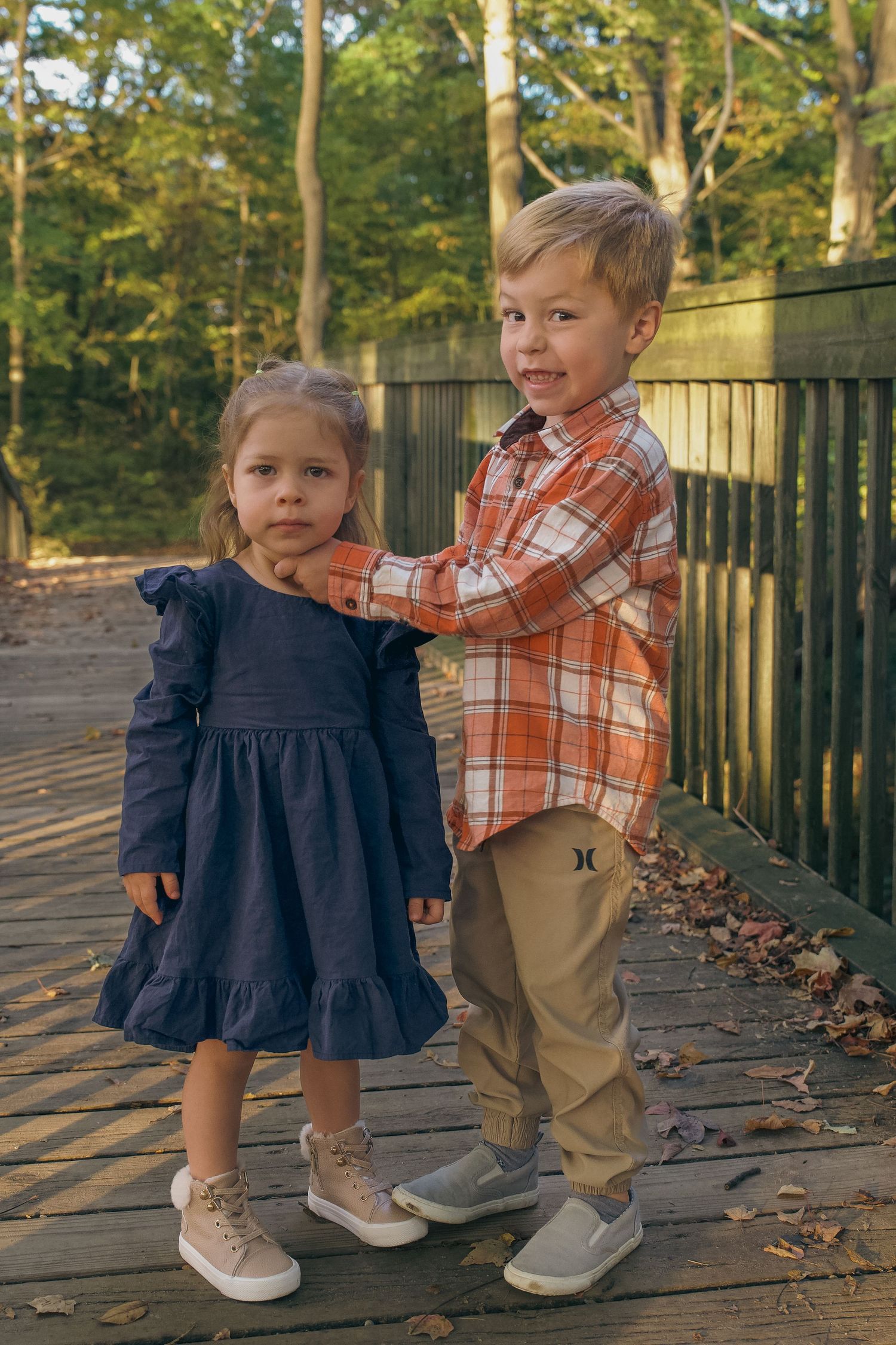Two young children in casual fall outfits play together on a wooden deck surrounded by autumn leaves.